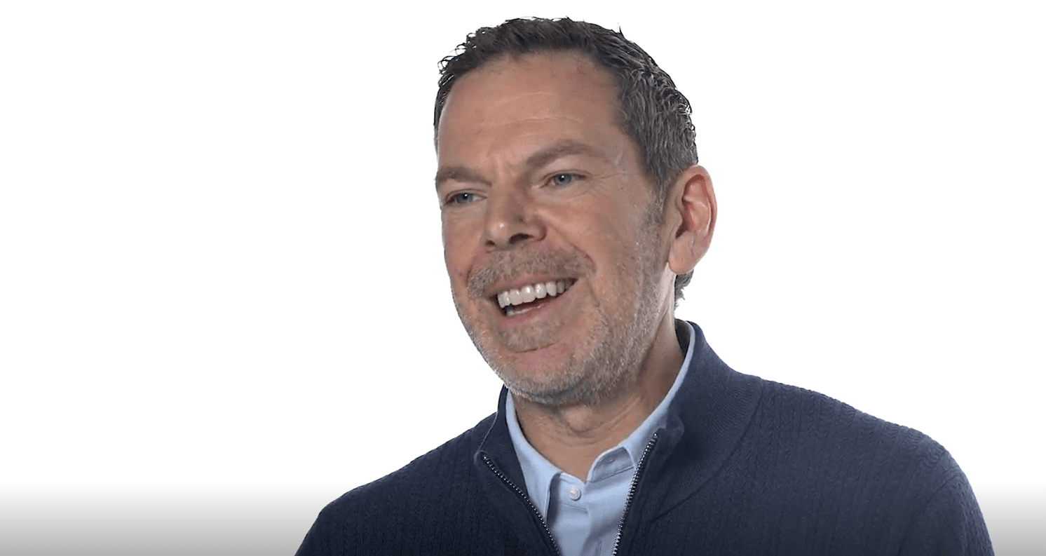 A man with dark hair and stubble, wearing a blue shirt and dark sweater, smiles broadly against a white background.