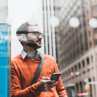 Man in glasses and an orange sweater holding a smartphone in a city.