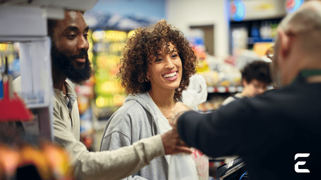 A smiling woman receives a bag from a cashier at a store checkout, with a man standing beside her.