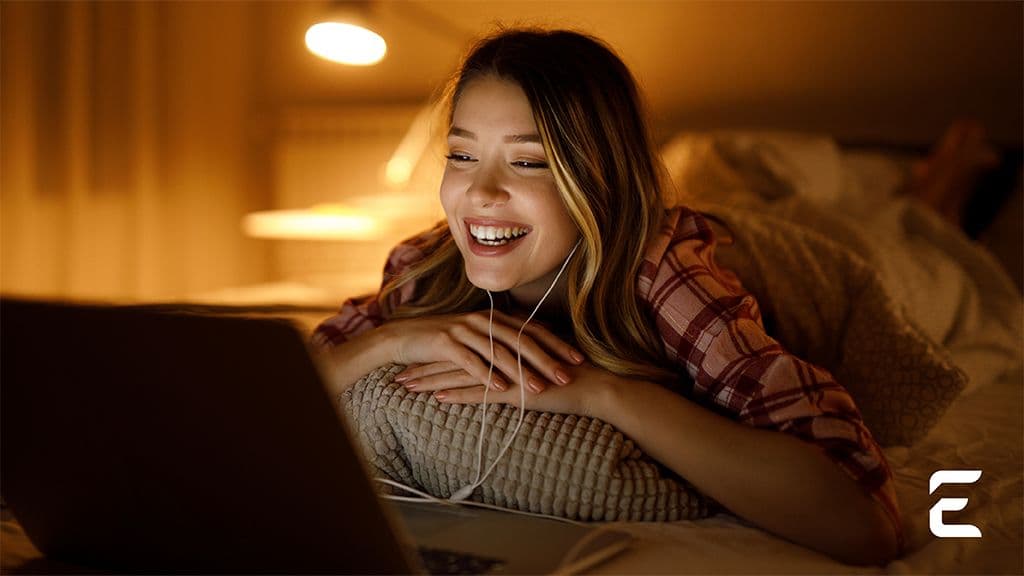 A warm-toned photo of a blonde woman smiling with corded headphones while she's watching something on her laptop