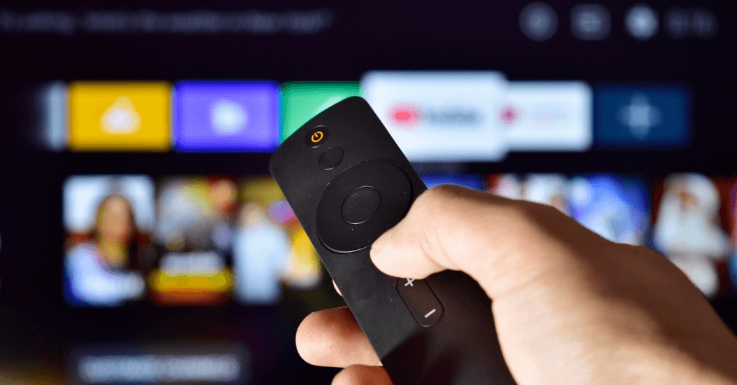 Someone holding a remote in front of a blurry background featuring icons of streaming platforms like YouTube and Hulu