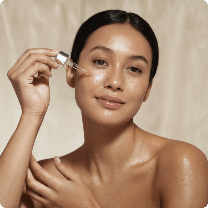 Smiling woman with glowing skin applying serum to her cheek with a dropper.