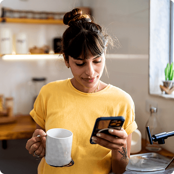 A young woman in a yellow shirt holds a white mug and looks at her smartphone in a bright kitchen.