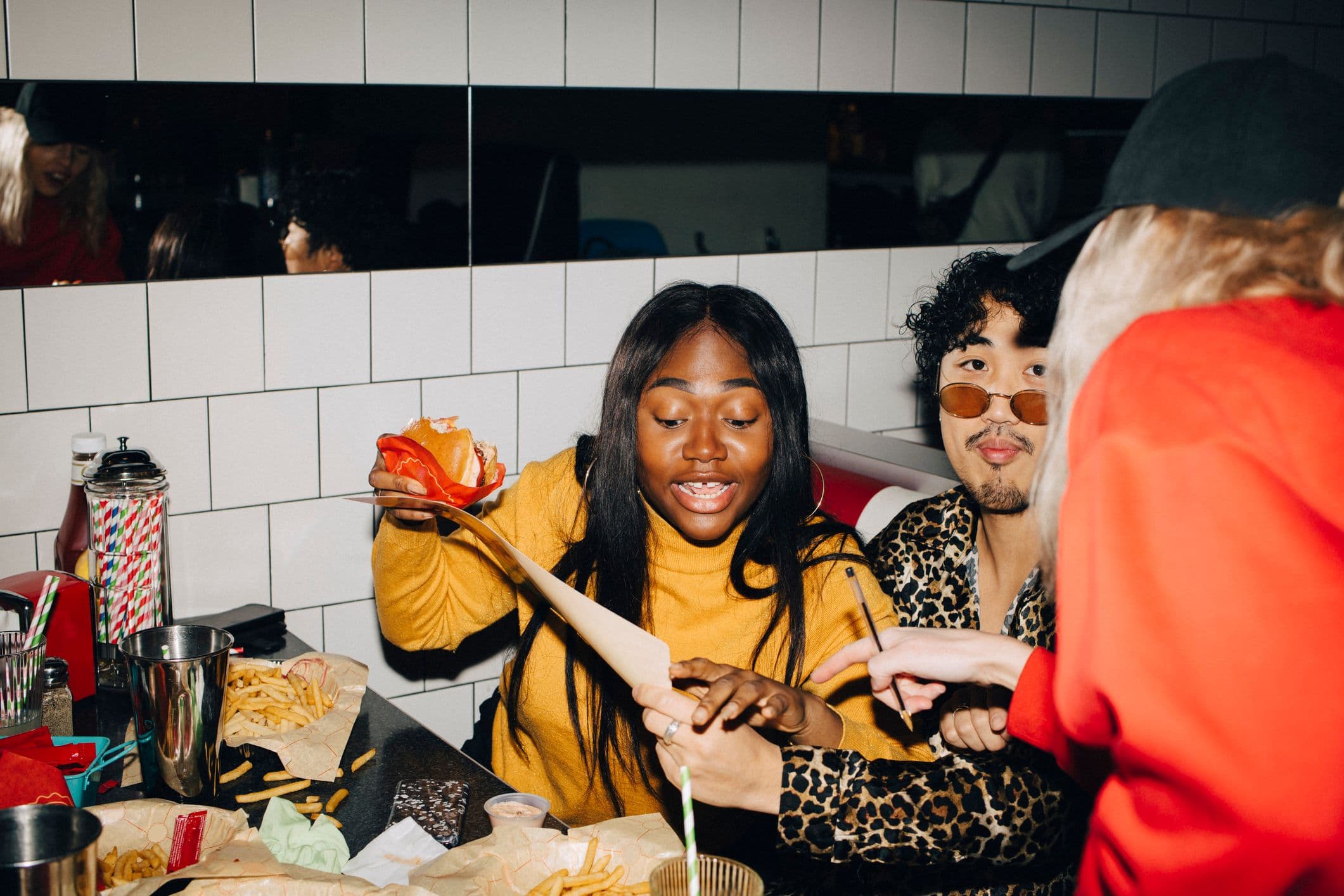 A young woman in a yellow sweater holds a burger while talking with friends at a diner table scattered with fries.