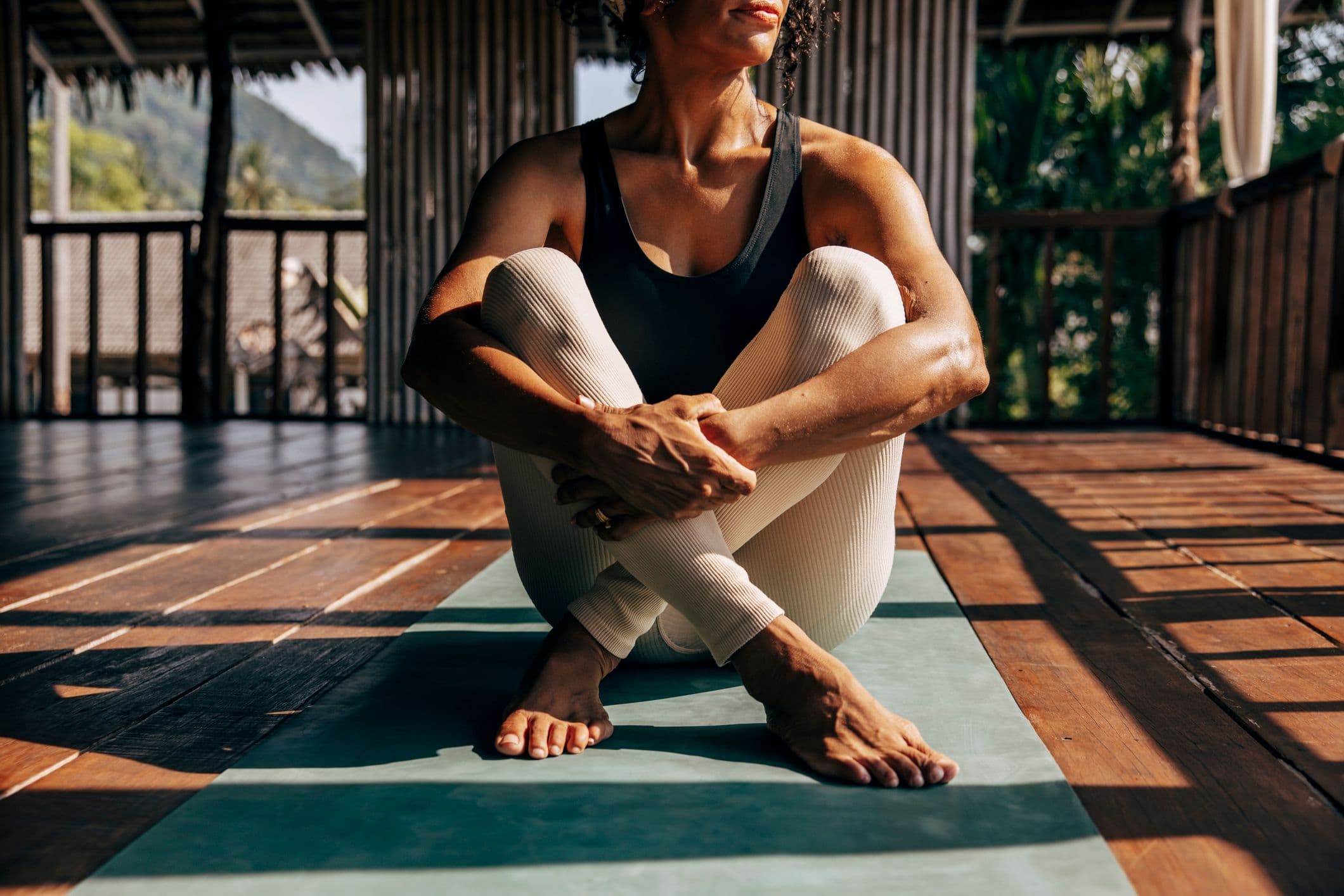 A woman sits cross-legged on a yoga mat on a wooden deck.