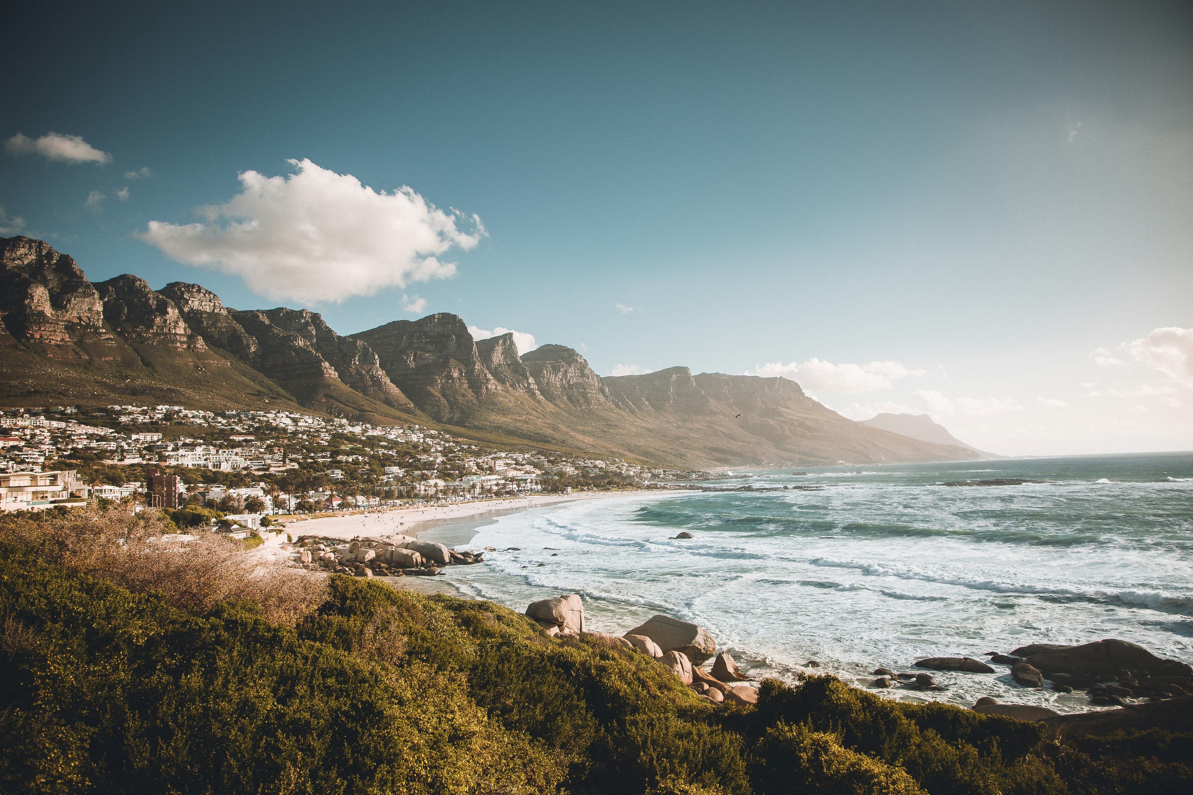 Coastal town and sandy beach with ocean waves, backed by rugged mountains under a blue sky.