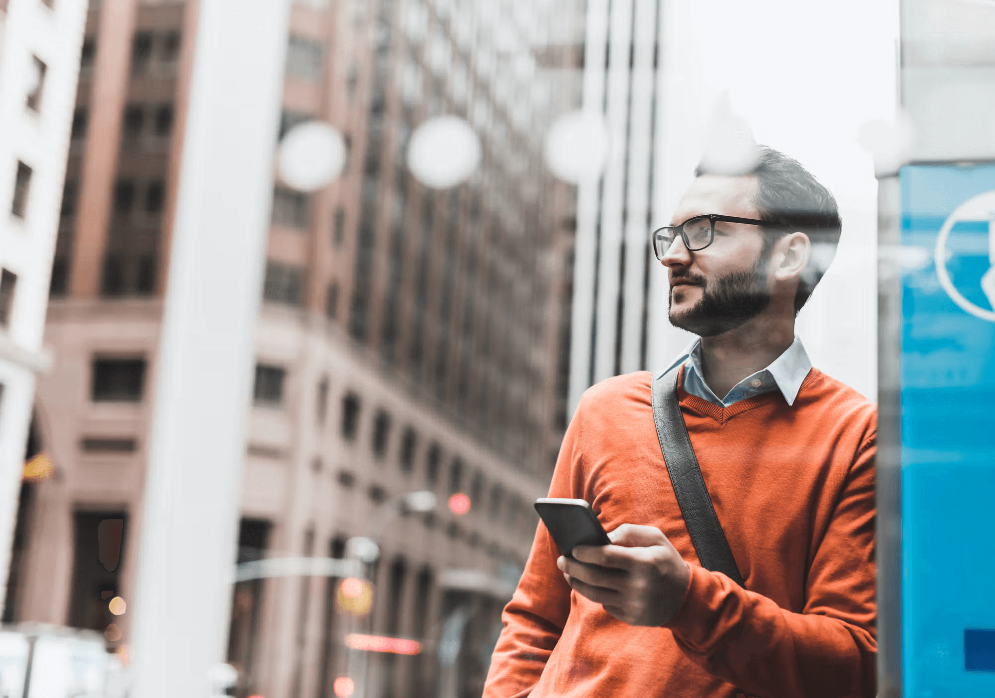 Man in an orange sweater, glasses, and beard holds a phone while looking right, with city buildings blurred in the background.
