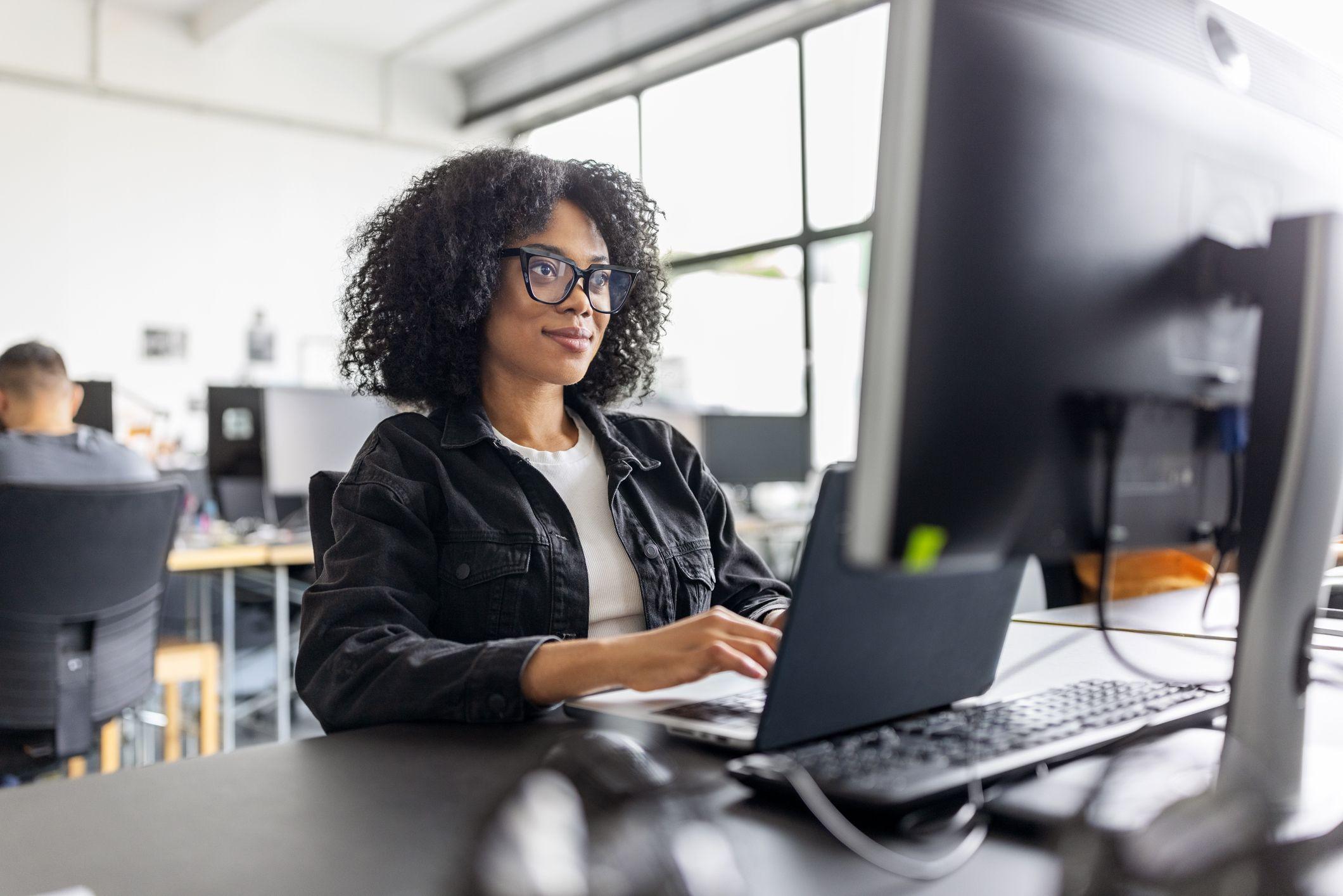 A smiling Black woman with curly hair and glasses types on a laptop at a desk in an office.