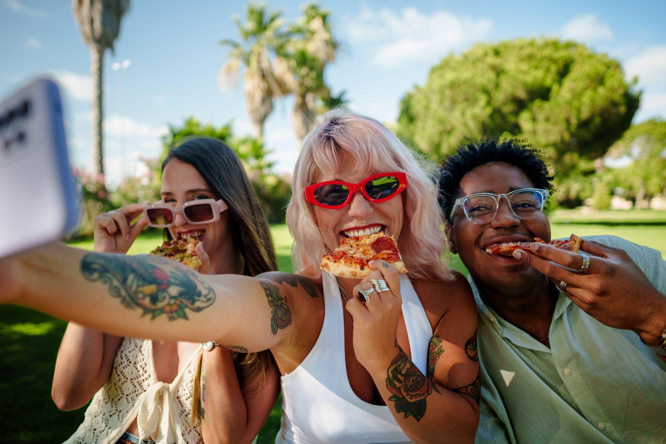 Three smiling friends taking a selfie while eating pizza outdoors.