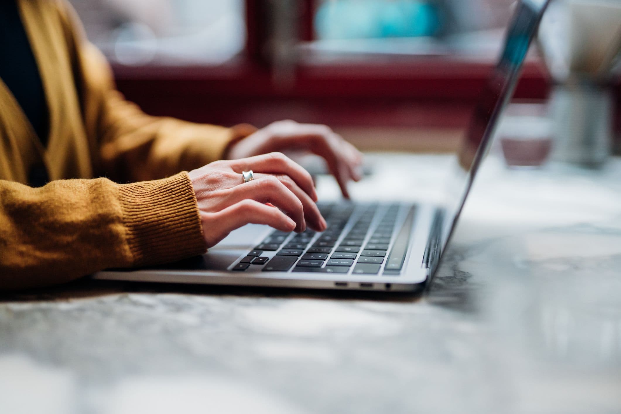 Hands in a mustard-yellow cardigan typing on on a laptop keyboard.