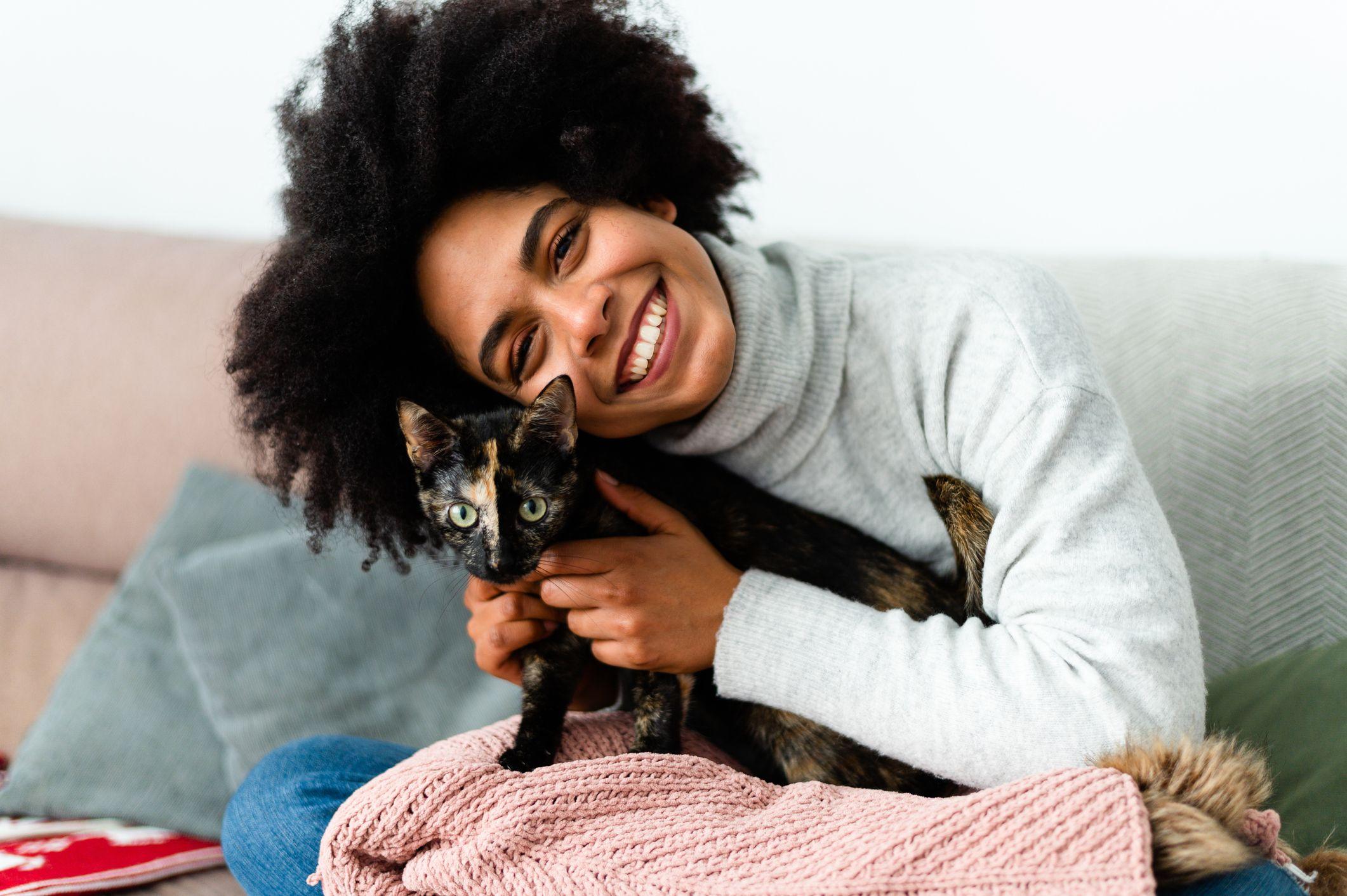 A smiling Black woman with an afro hugs a tortoiseshell cat.
