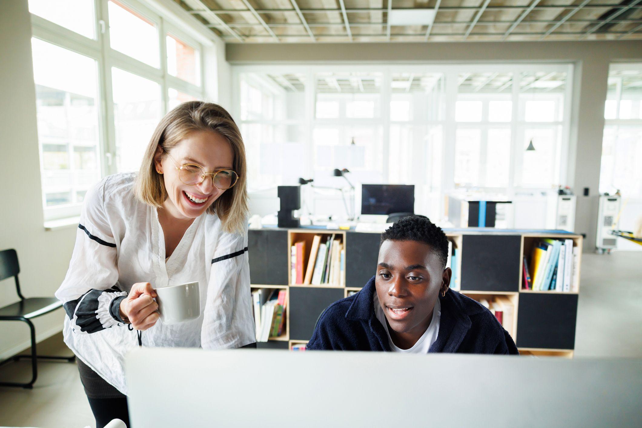 A laughing woman and a focused man look at a computer in an office.